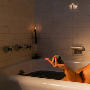 Black Magic CBD Bath Bomb being held in a hand with red nails over a white bathtub with lit candles. 